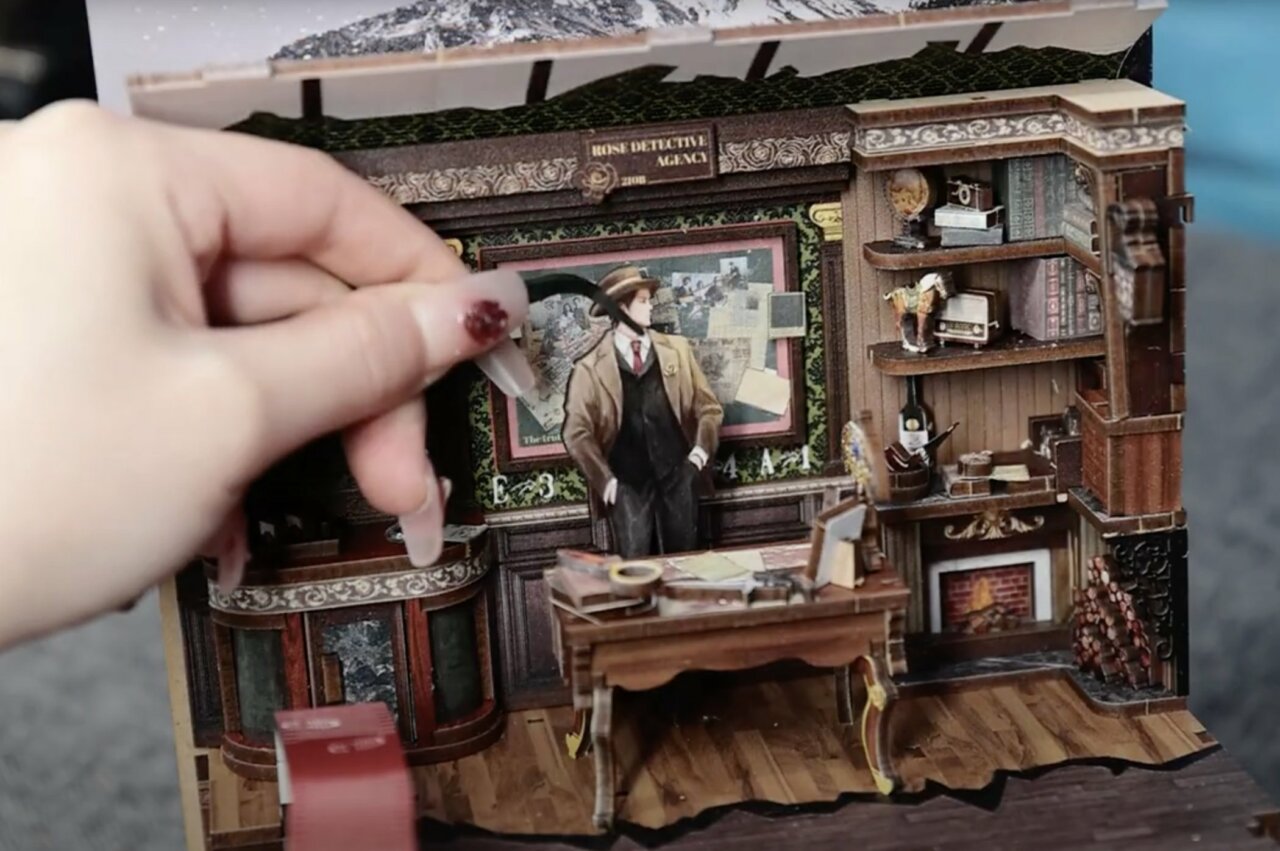 Manos ensamblando un book nook con una escena en miniatura de despacho antiguo al estilo detective, con figuras, muebles y detalles victorianos, representando el proceso artesanal de construcción de un book nook