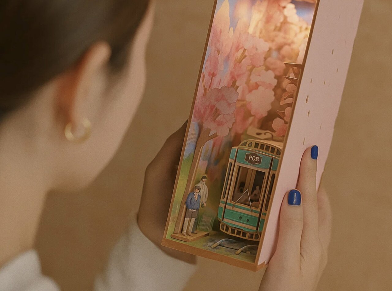 Persona sosteniendo un book nook sakura con un tranvía en miniatura rodeado de cerezos en flor, iluminado con luz cálida, símbolo de calma, belleza y detalle artesanal japonés