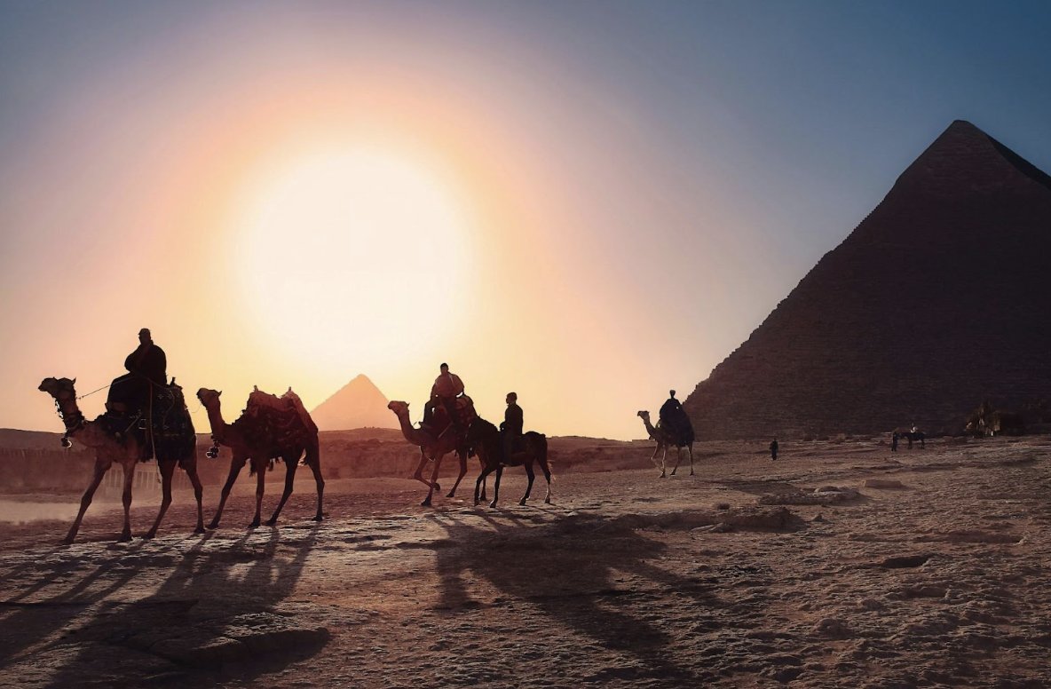 Camellos y personas en el desierto junto a las pirámides de Egipto al atardecer.