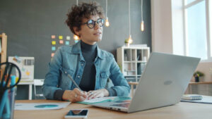 Mujer escribiendo en un cuaderno frente a una computadora portátil