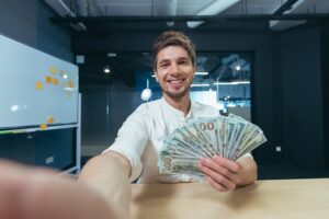 Hombre sonriente mostrando billetes de cien d&oacute;lares en una oficina moderna